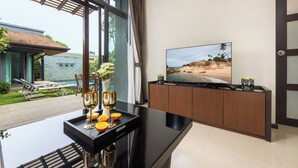 A modern living room with a large TV, a dining table with a tray of glasses and oranges, and a view of a beach through large windows.