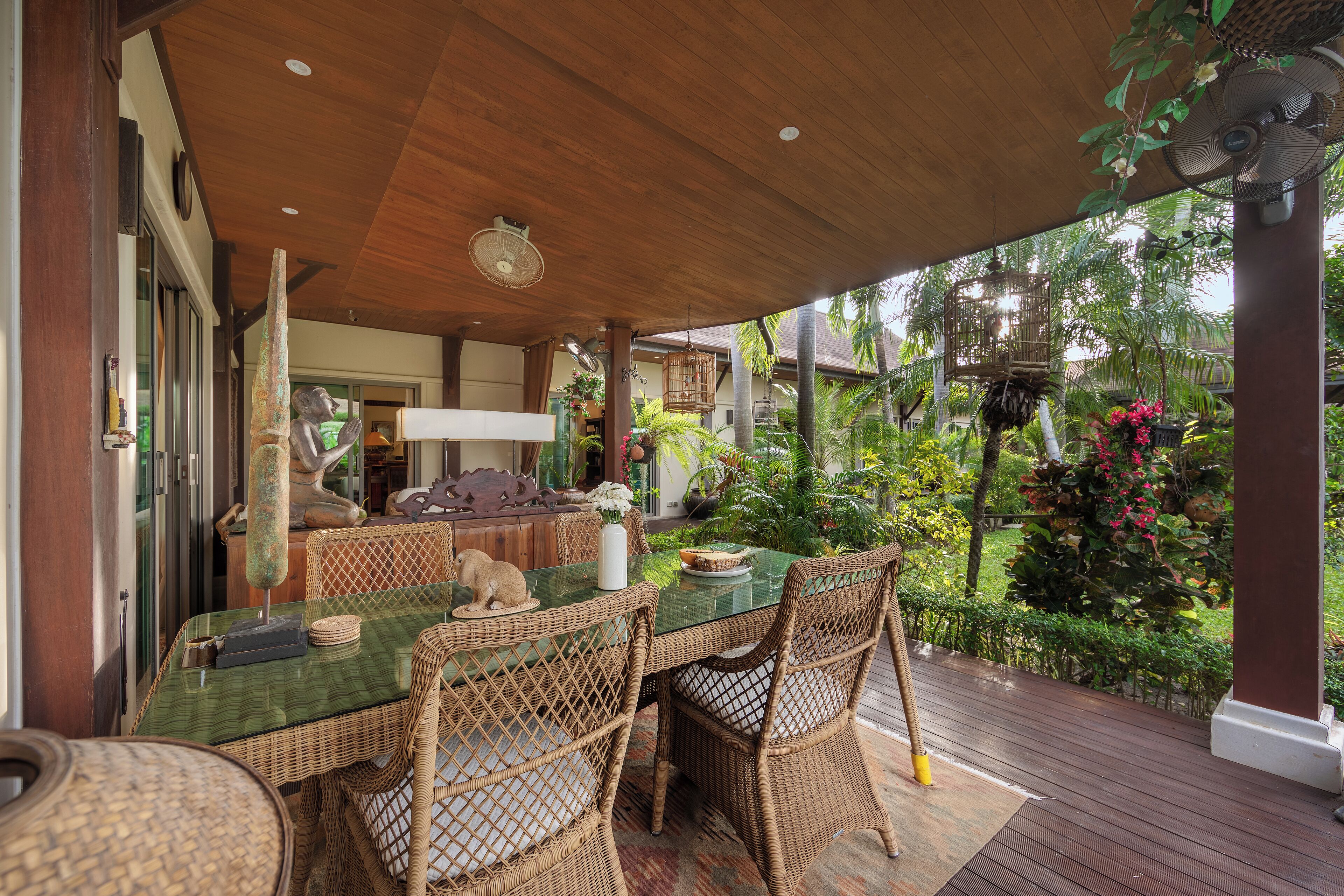 An outdoor dining area with wicker furniture and a glass table, surrounded by lush greenery and a wooden deck.