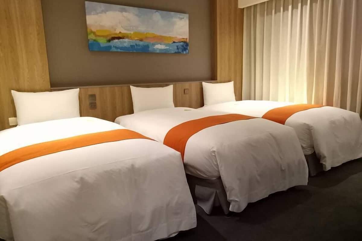Ein Hotelzimmer mit drei Betten, jeweils mit weißer Bettwäsche und orangenen Akzenten. Darüber hängt ein Gemälde an der Wand.