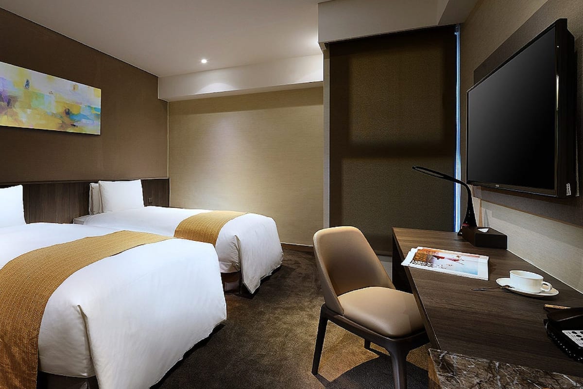 Ein Hotelzimmer mit zwei Betten, einem Fernseher, einem Schreibtisch mit Stuhl und einem Bild an der Wand.