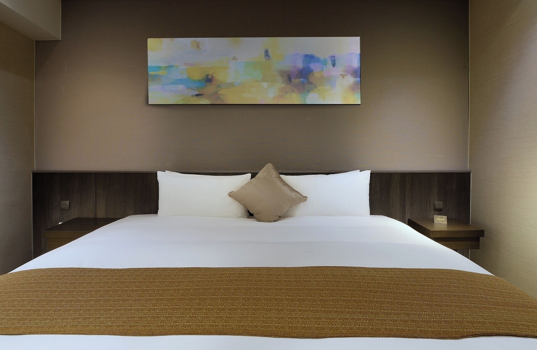 A hotel room with a large bed, a beige throw blanket, and a colorful abstract painting on the wall.