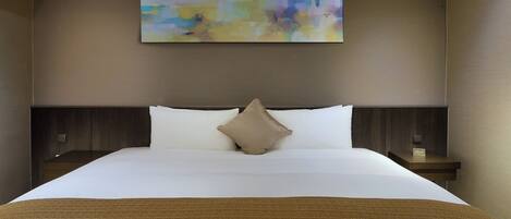 A hotel room with a large bed, a beige throw blanket, and a colorful abstract painting on the wall.