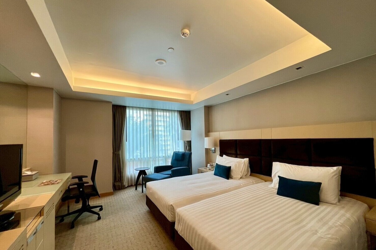 Una habitación de hotel con dos camas, un escritorio con computadora, una silla y una ventana con cortinas.
