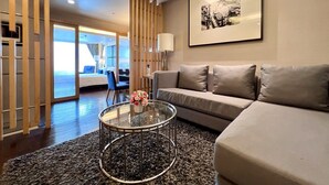 A modern living room with a sofa, a glass coffee table with flowers, a framed picture on the wall, and a view of the sea through large windows. - Sora Resort & Suites Sukhumvit (Bangkok)