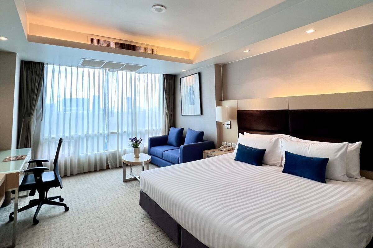 Habitación de hotel con una cama grande, un escritorio, una silla y un sofá. Hay una ventana con cortinas y vistas a la ciudad.