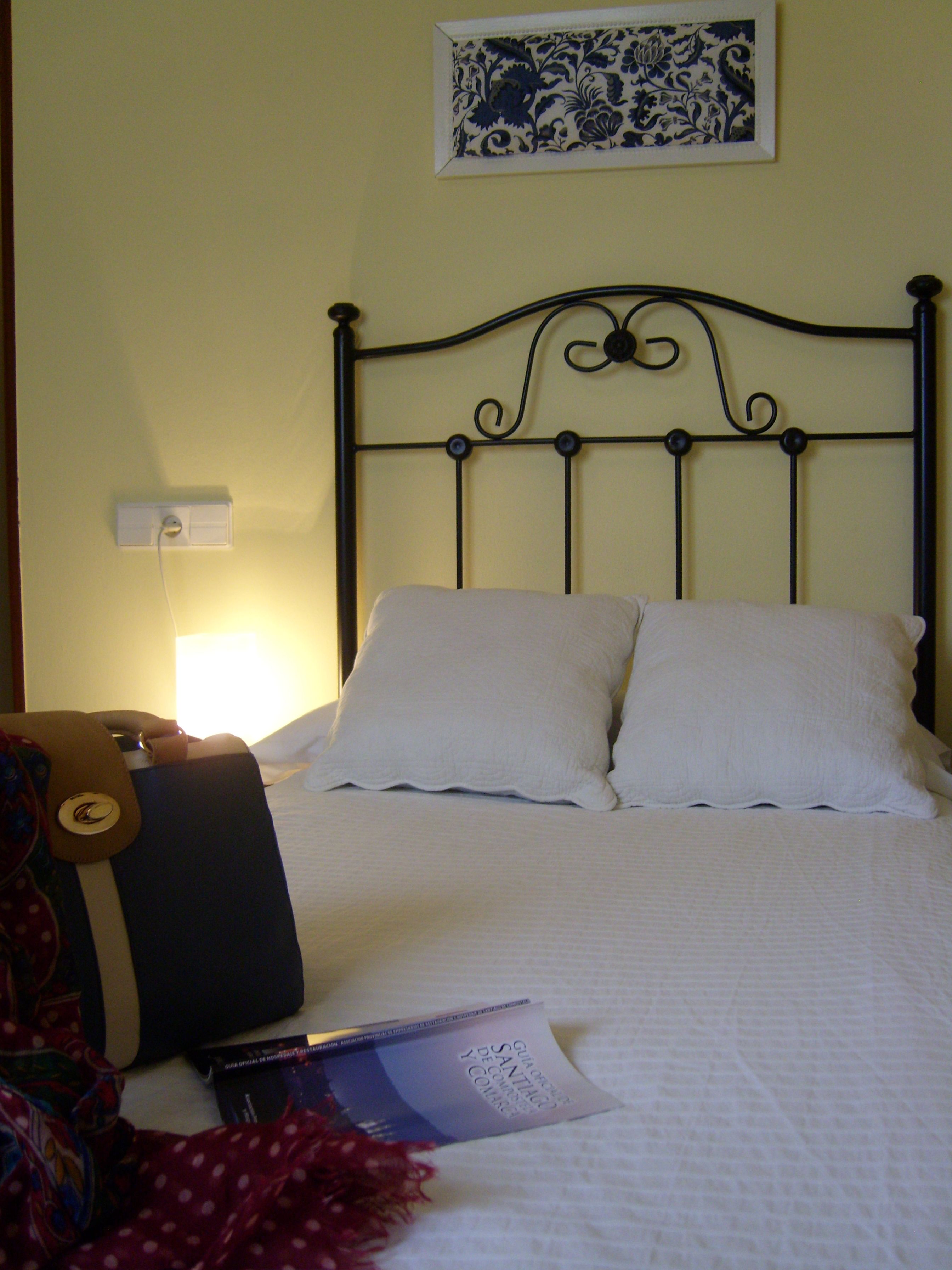 Een bed met witte kussens en een zwart metalen hoofdbord. Boven het bed hangt een foto in een lijst aan een witte muur.