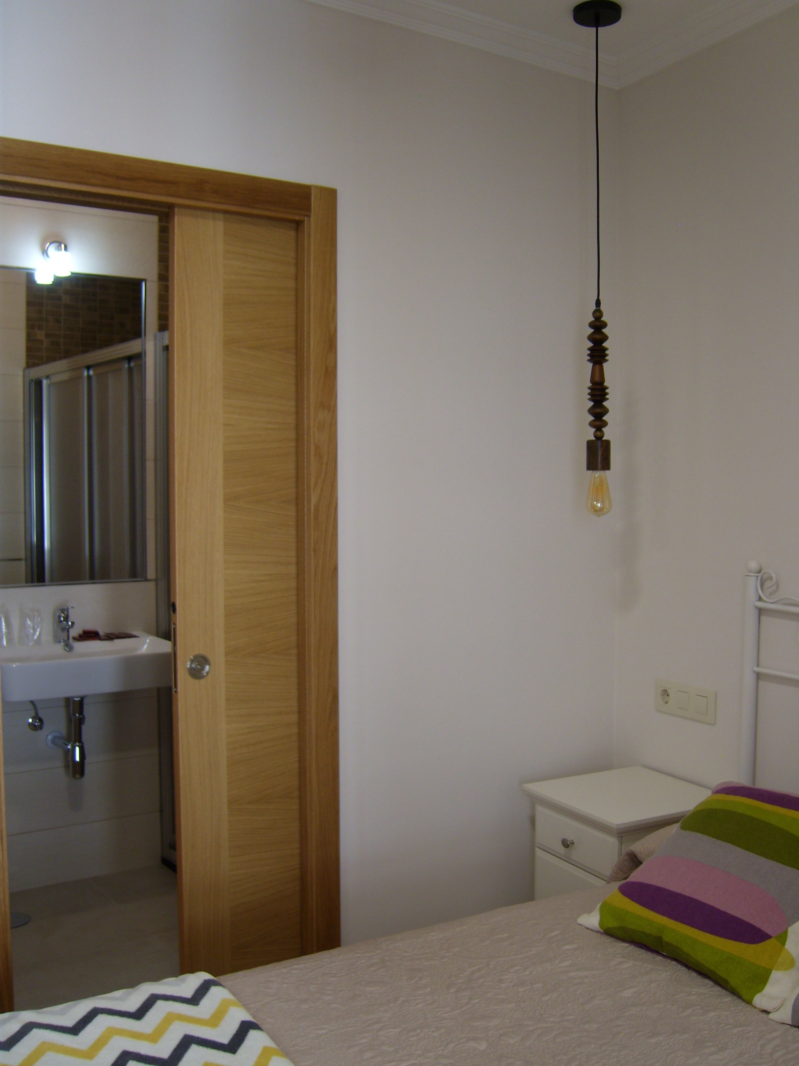Un dormitori amb un llit, una taula de nit, una porta de fusta i un bany amb un lavabo i un mirall.