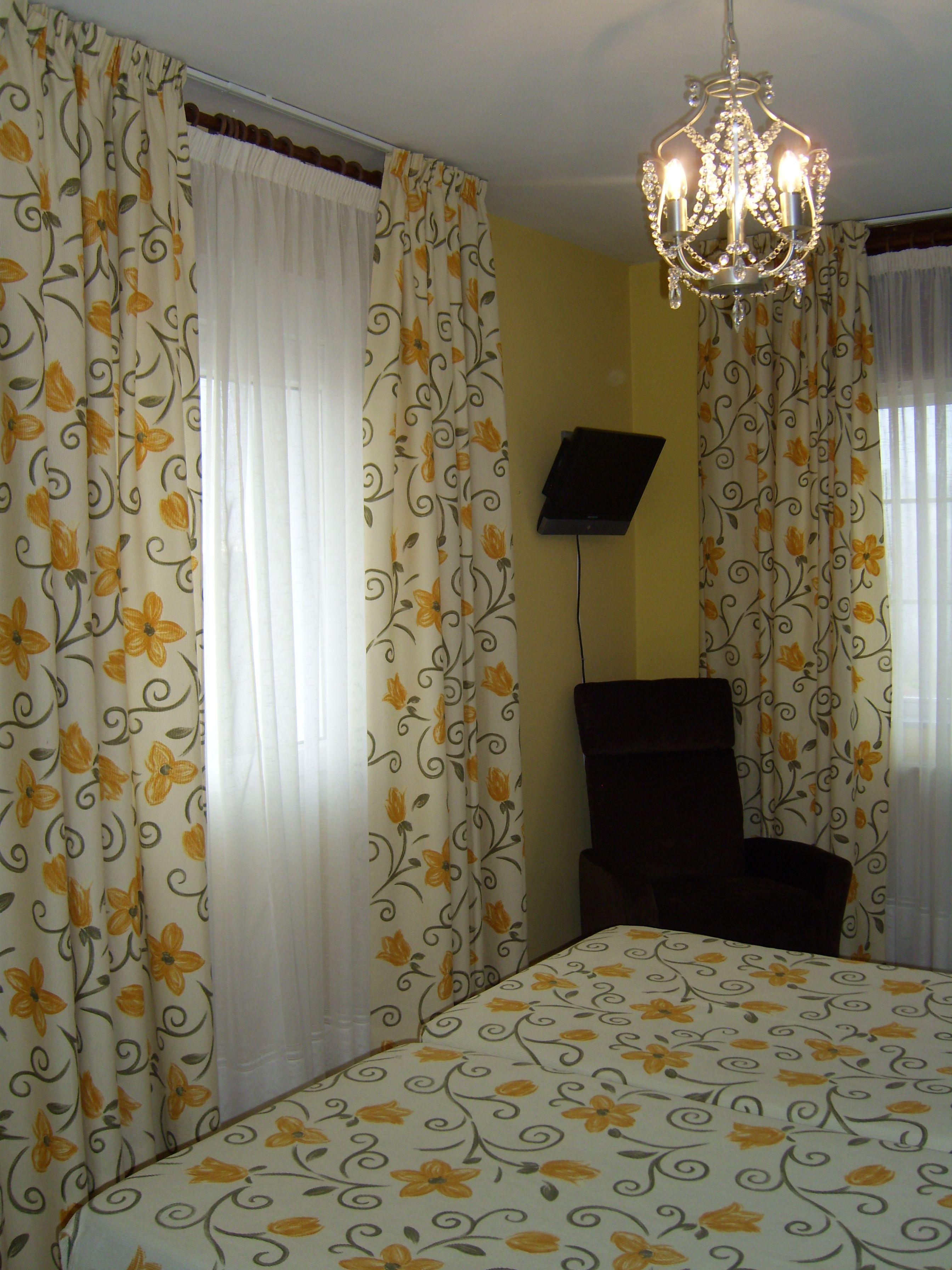Habitación de hotel con cortinas florales, una cama con colcha estampada, un candelabro y un televisor en la pared.