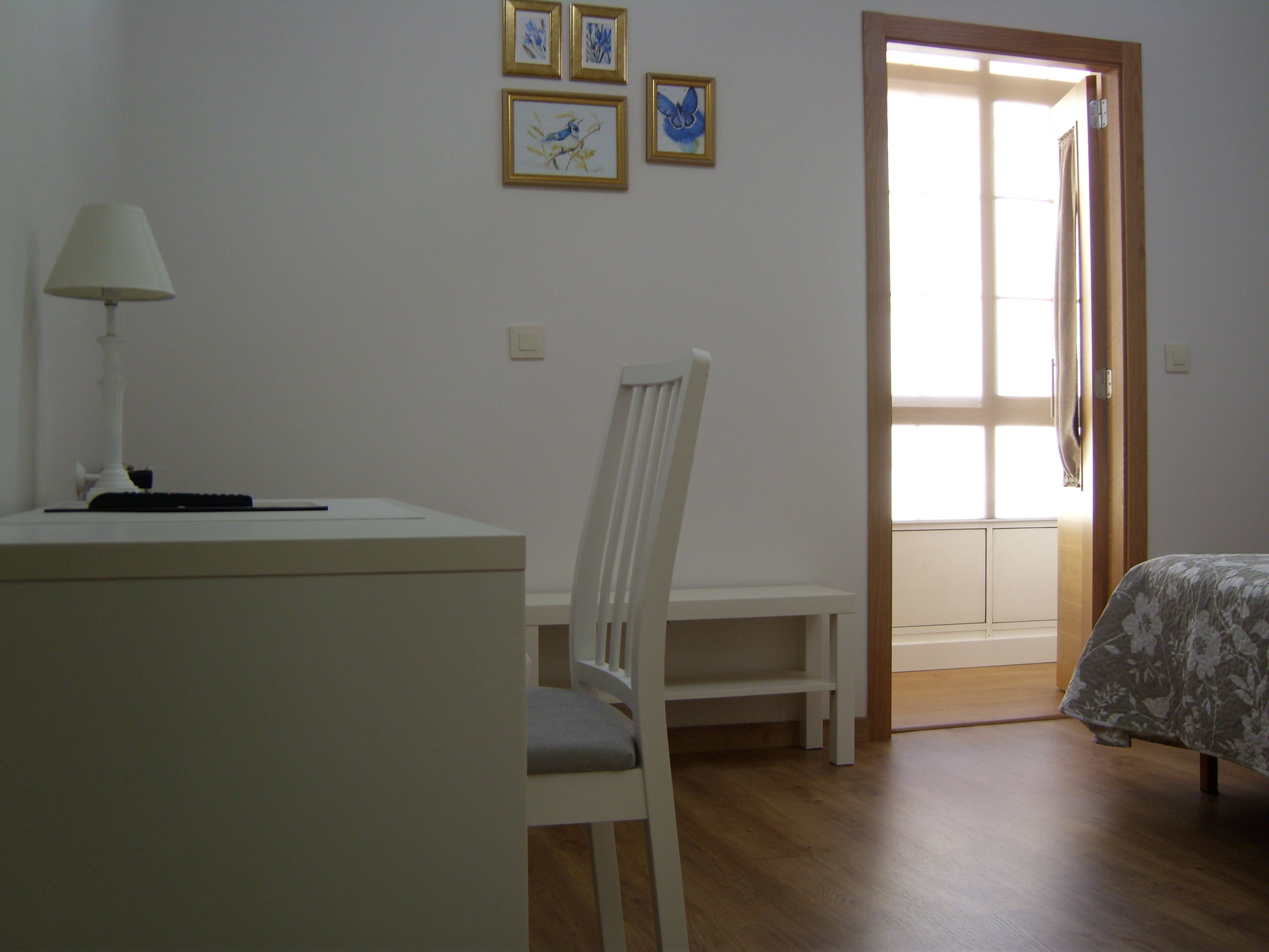 A room with a desk, chair, bed, and a door leading to another room.