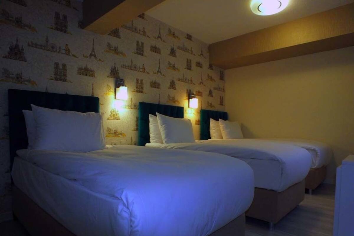 A hotel room with two beds, each with white bedding and headboards. There are wall-mounted lights above the beds, and the walls are decorated with a pattern of cityscapes.
