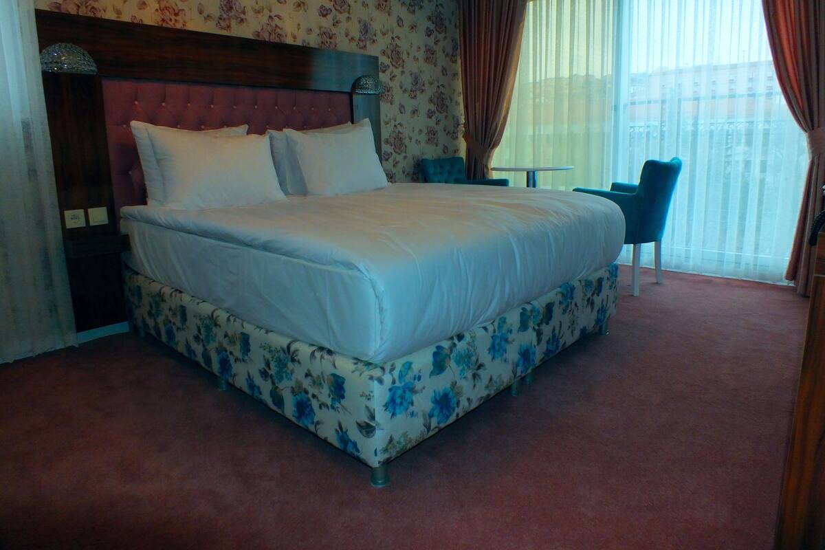 A hotel room with a large bed, floral wallpaper, a wooden headboard, a small table, and a chair.
