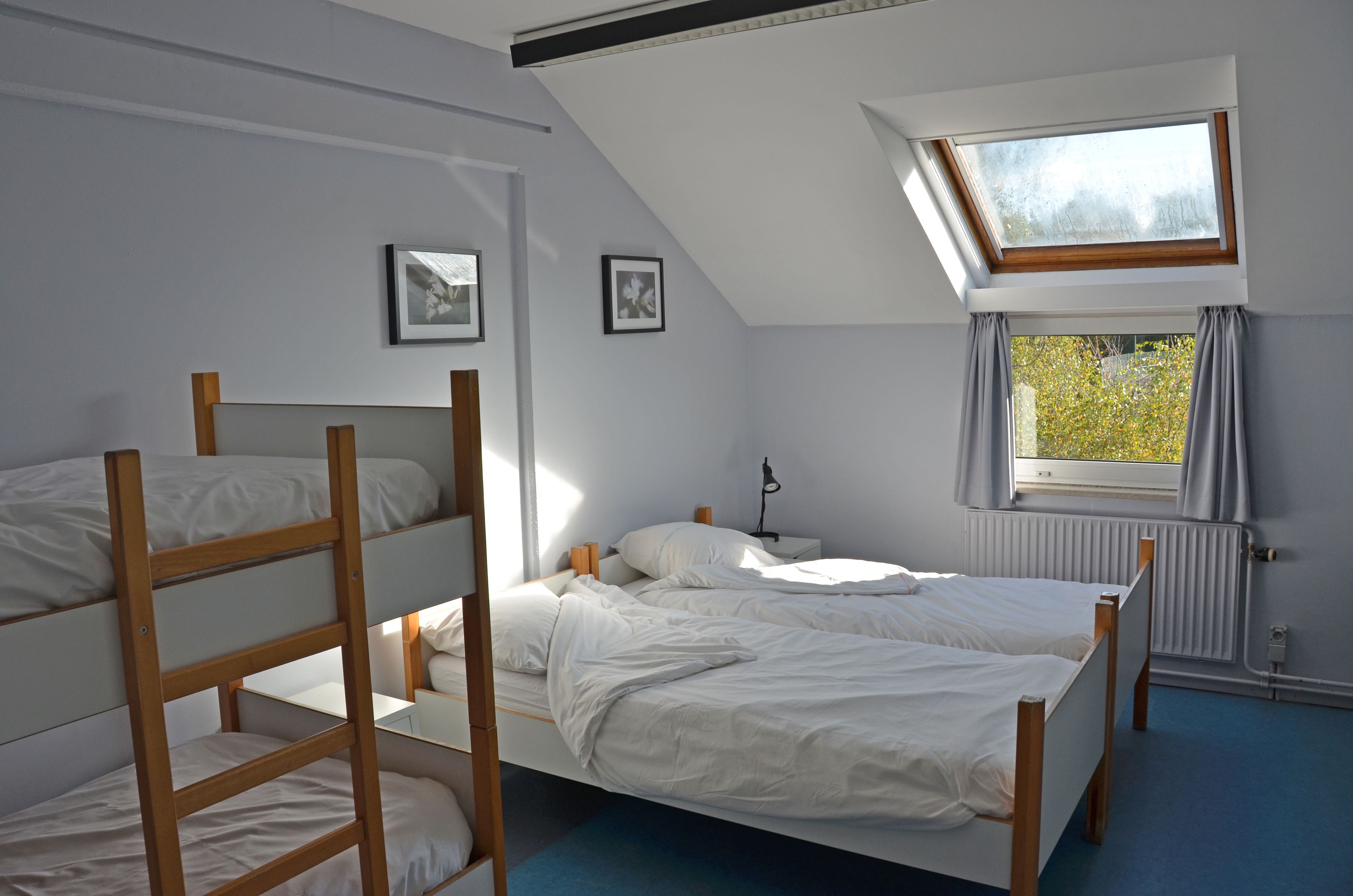 Een studentenkamer met stapelbedden, een dakraam, een raam met gordijnen en een radiator.