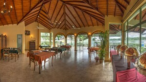 Un amplio comedor interior con techos altos de madera, varias mesas y grandes ventanales con vistas a un paisaje exuberante.