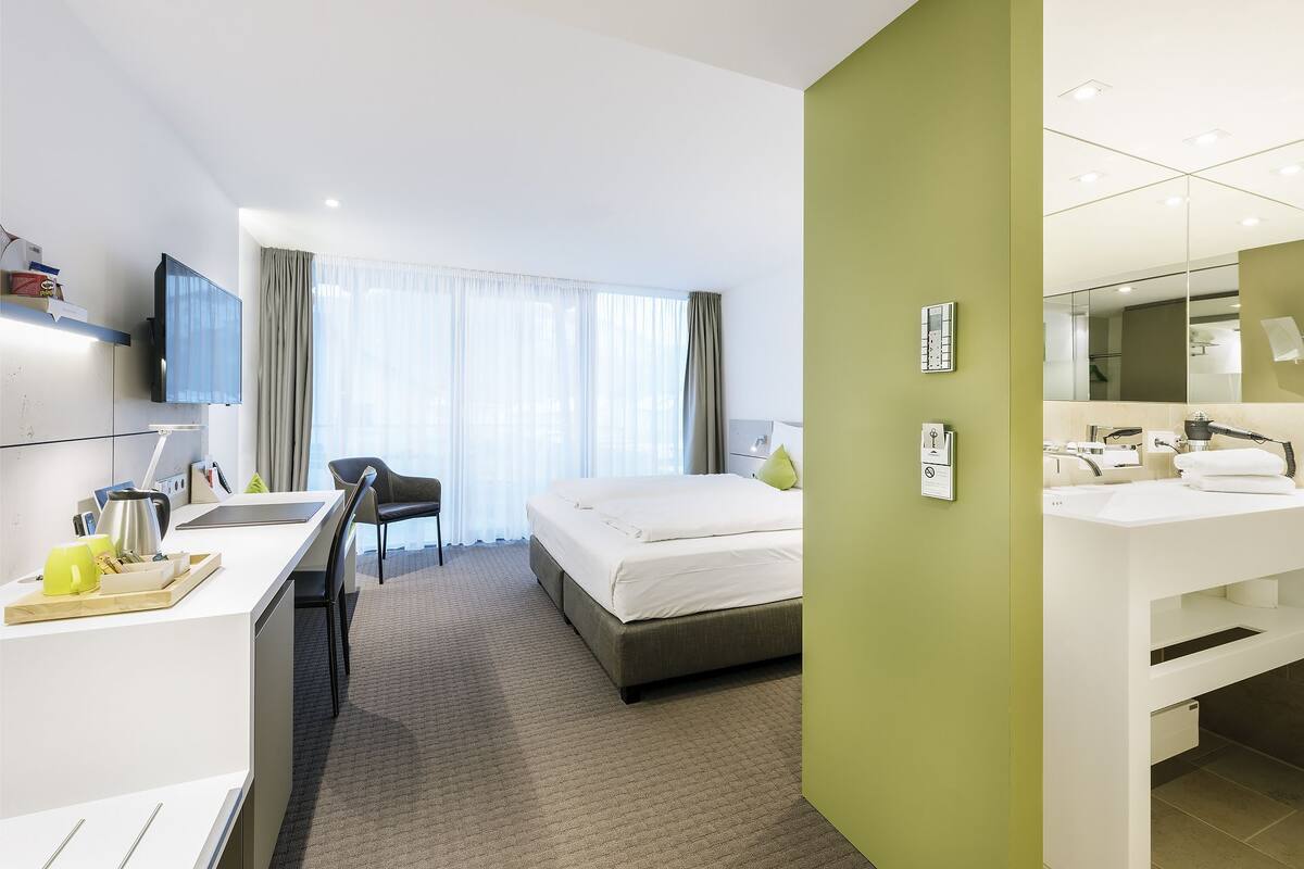 A hotel room with a bed, desk, chair, and a bathroom with a sink and mirror.