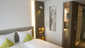 A hotel room with a bed, pillows, a wall-mounted light, a small shelf with a plant, and a door leading to another room. - HGS3 Konzept-Hotel (Schelklingen)