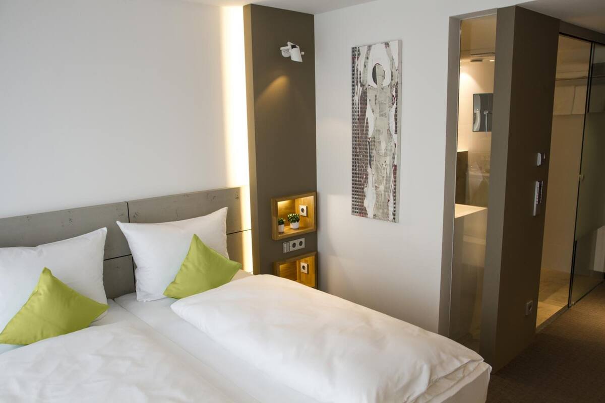 Habitación de hotel con cama, almohadas, una lámpara de pared, un pequeño estante con una planta y una puerta que conduce a otra habitación.