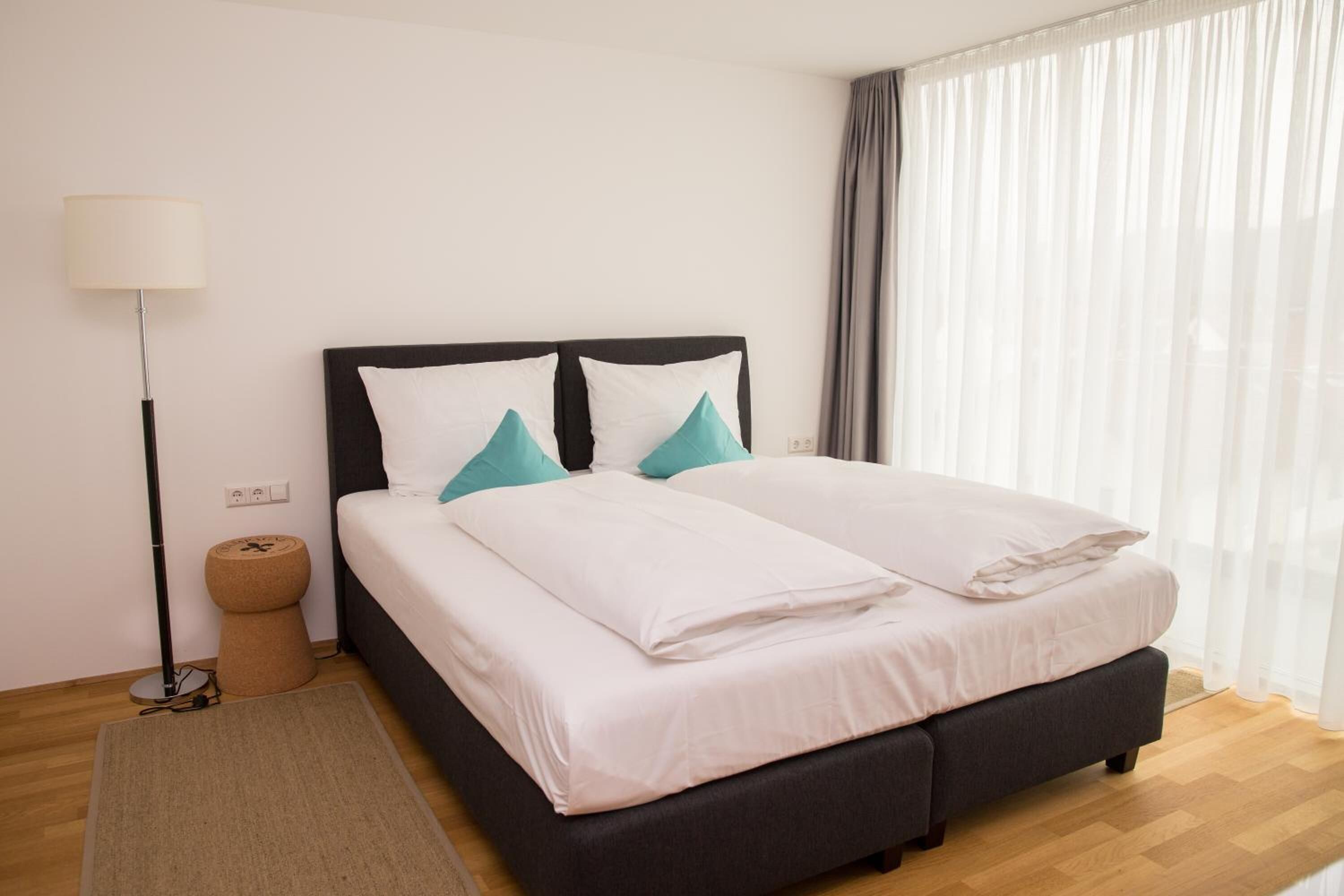 A neatly made bed with white and teal pillows, a black headboard, and a light-colored rug on a wooden floor.