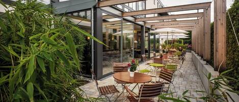 An outdoor patio with tables and chairs, surrounded by greenery and a glass structure.