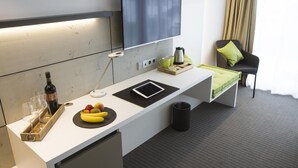 A modern hotel room with a white desk, a flat-screen TV, a small seating area with a chair and a cushion, and a window with curtains. - HGS3 Konzept-Hotel (Schelklingen)