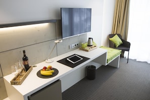 Habitación de hotel moderna con escritorio blanco, televisor de pantalla plana, una pequeña zona de estar con una silla y un cojín, y una ventana con cortinas.
