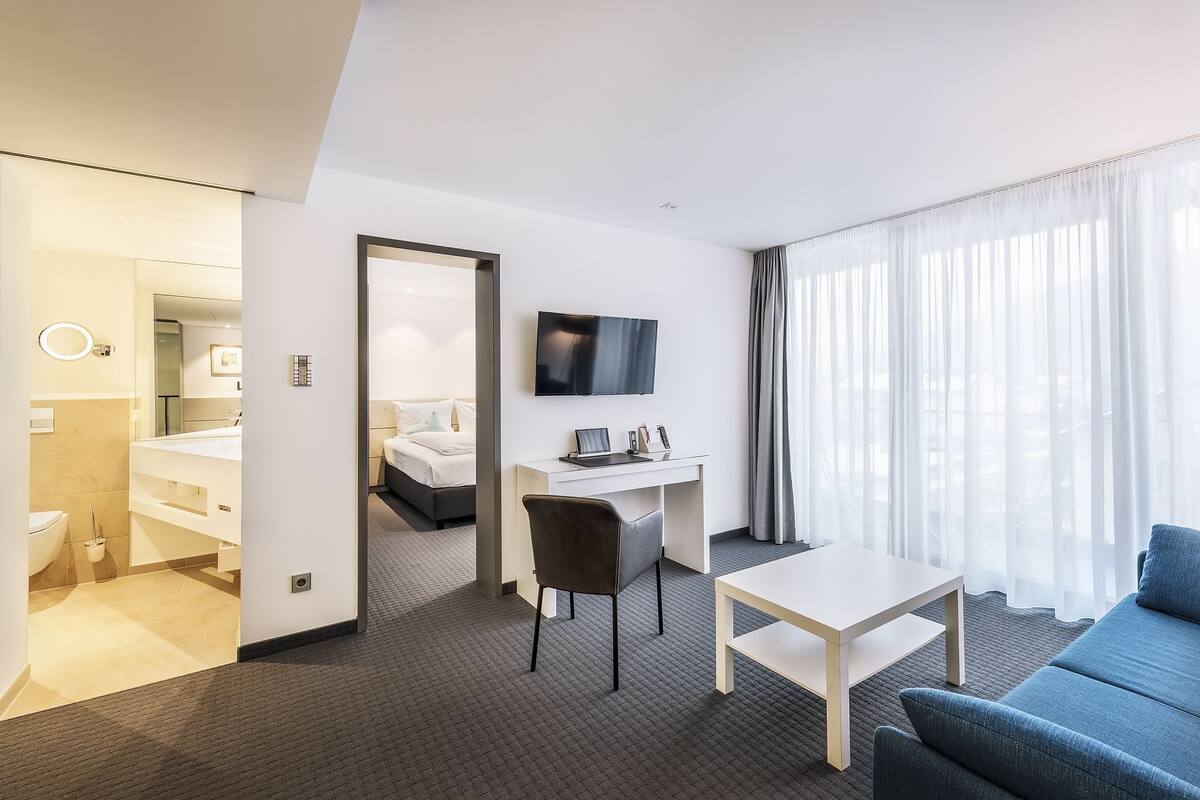 A hotel room with a bed, a desk with a chair, a TV, a sofa, and a bathroom visible through an open door.