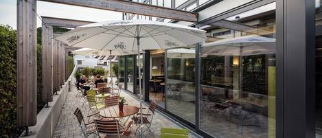 Un café al aire libre con mesas y sillas, grandes sombrillas y un muro de cristal que refleja el interior.