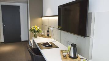 Une cuisine moderne avec un plan de travail blanc, un grand téléviseur à écran plat fixé au-dessus, un vase avec des fleurs, une tablette et une bouilloire posés sur une planche en bois.