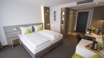A hotel room with a bed, bedside table, desk, chair, and a small table with a kettle and cup.