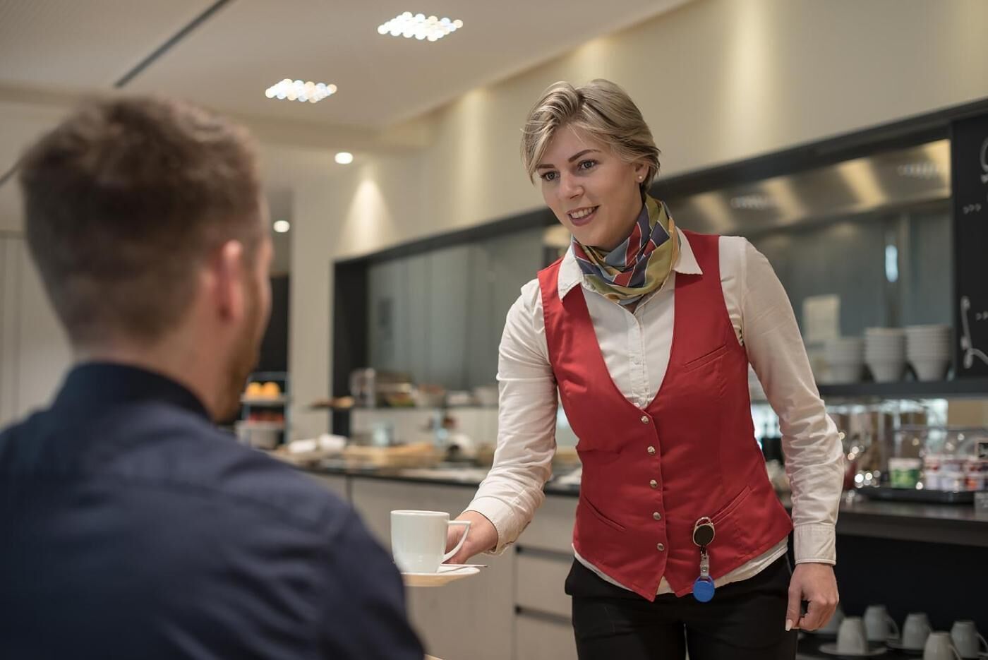 Un cameriere con un grembiule rosso e una camicia bianca serve un cliente in un moderno caffè, con un bancone e diversi cibi in vista.