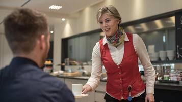 Un cameriere con un grembiule rosso e una camicia bianca serve un cliente in un moderno caffè, con un bancone e diversi cibi in vista.