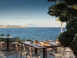 Zona pranzo all'aperto con tavoli e sedie, affacciata su una distesa d'acqua con una costa e montagne in lontananza.