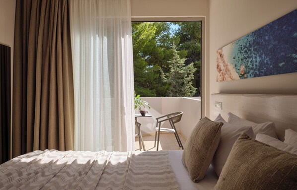 A bedroom with a bed, curtains, a view of trees, a small table with a plant, and a painting on the wall.