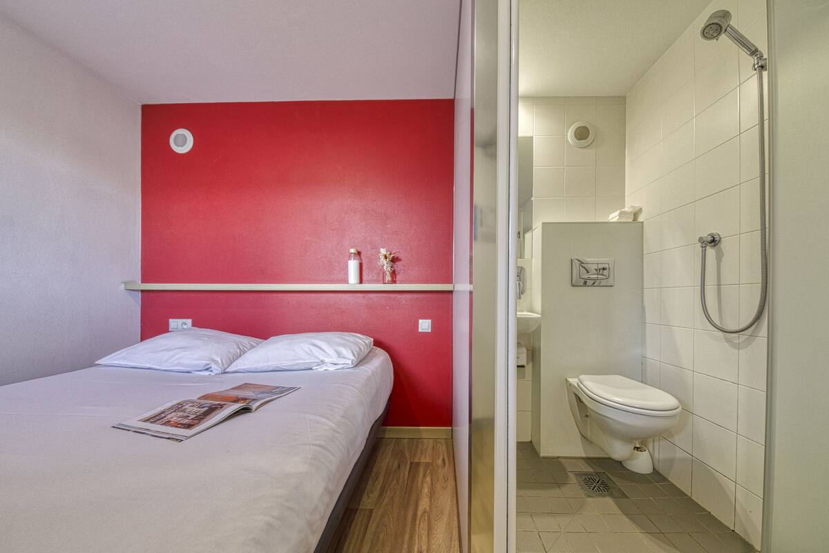 A hotel room with a bed, a red accent wall, a shower, and a toilet.