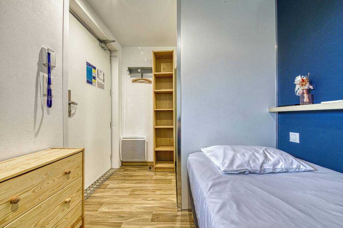 A bedroom with a bed, a wooden dresser, a shelf with a vase, and a blue wall.