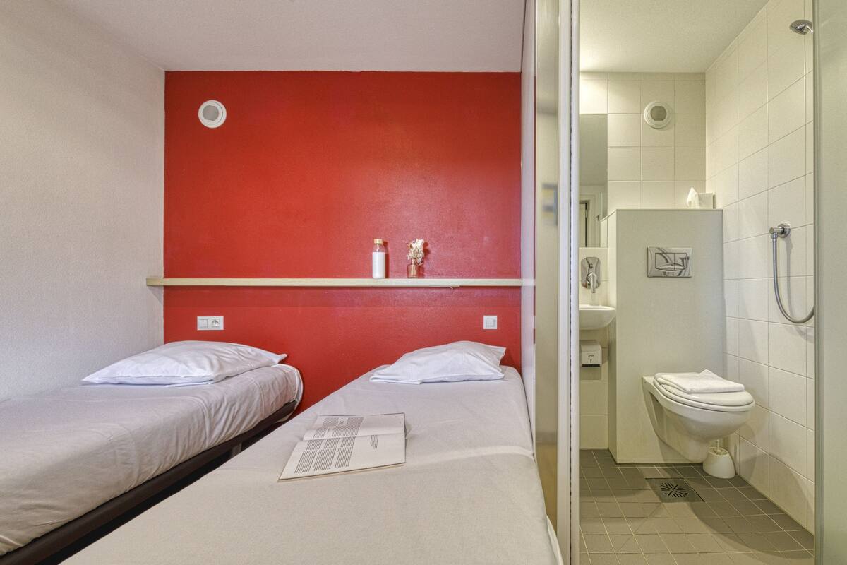 A small, compact hotel room with two single beds, a red accent wall, a small shelf with toiletries, and a separate bathroom with a toilet and shower.
