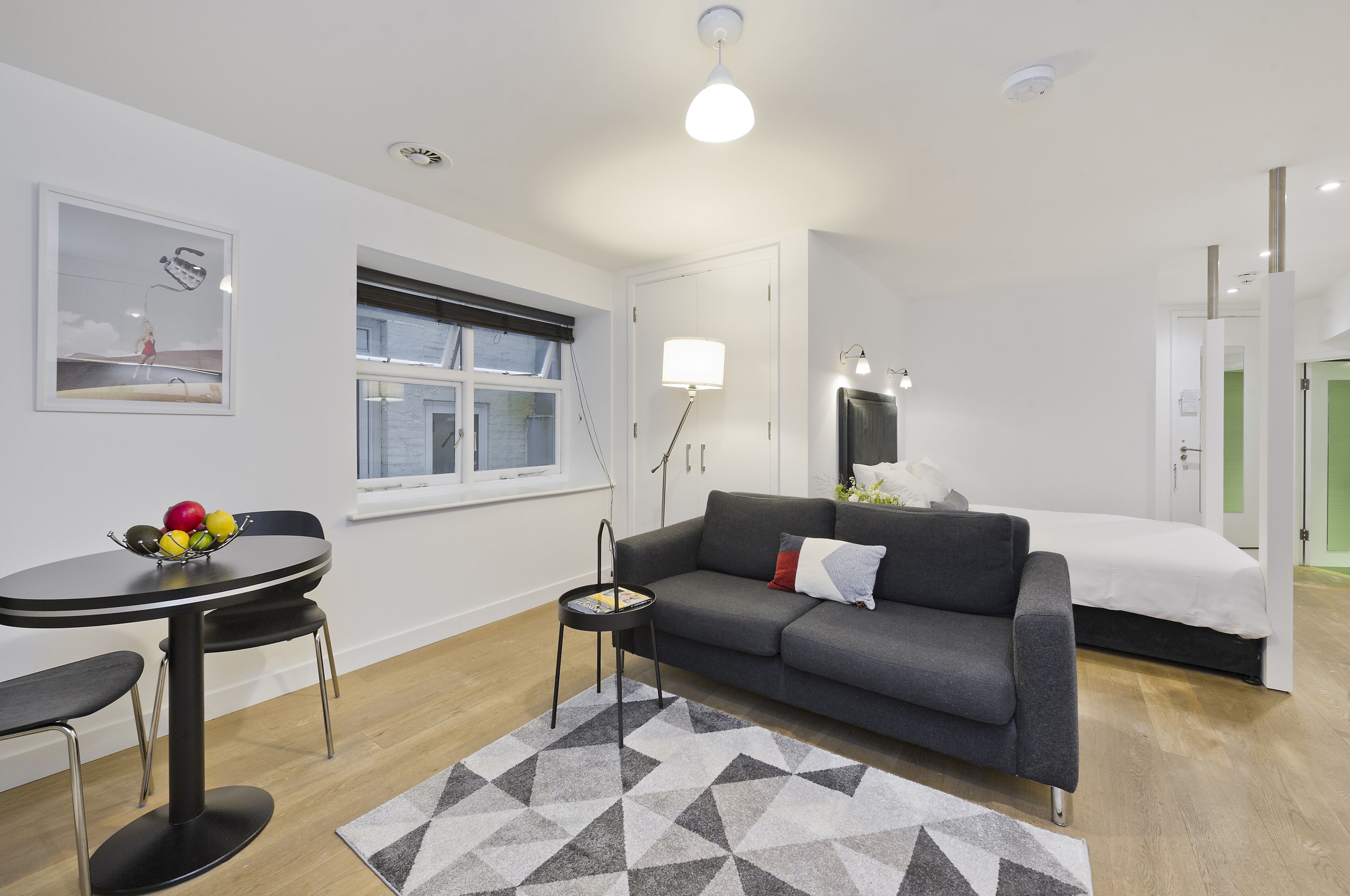 A modern, well-lit room with a grey sofa, a small round table with a fruit bowl, a black chair, a geometric rug, and a bed with a white mattress.