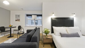 Una habitación de hotel moderna con una cama grande, un sofá, una mesita con sillas, una ventana con persianas y una lámpara fijada a la pared.
