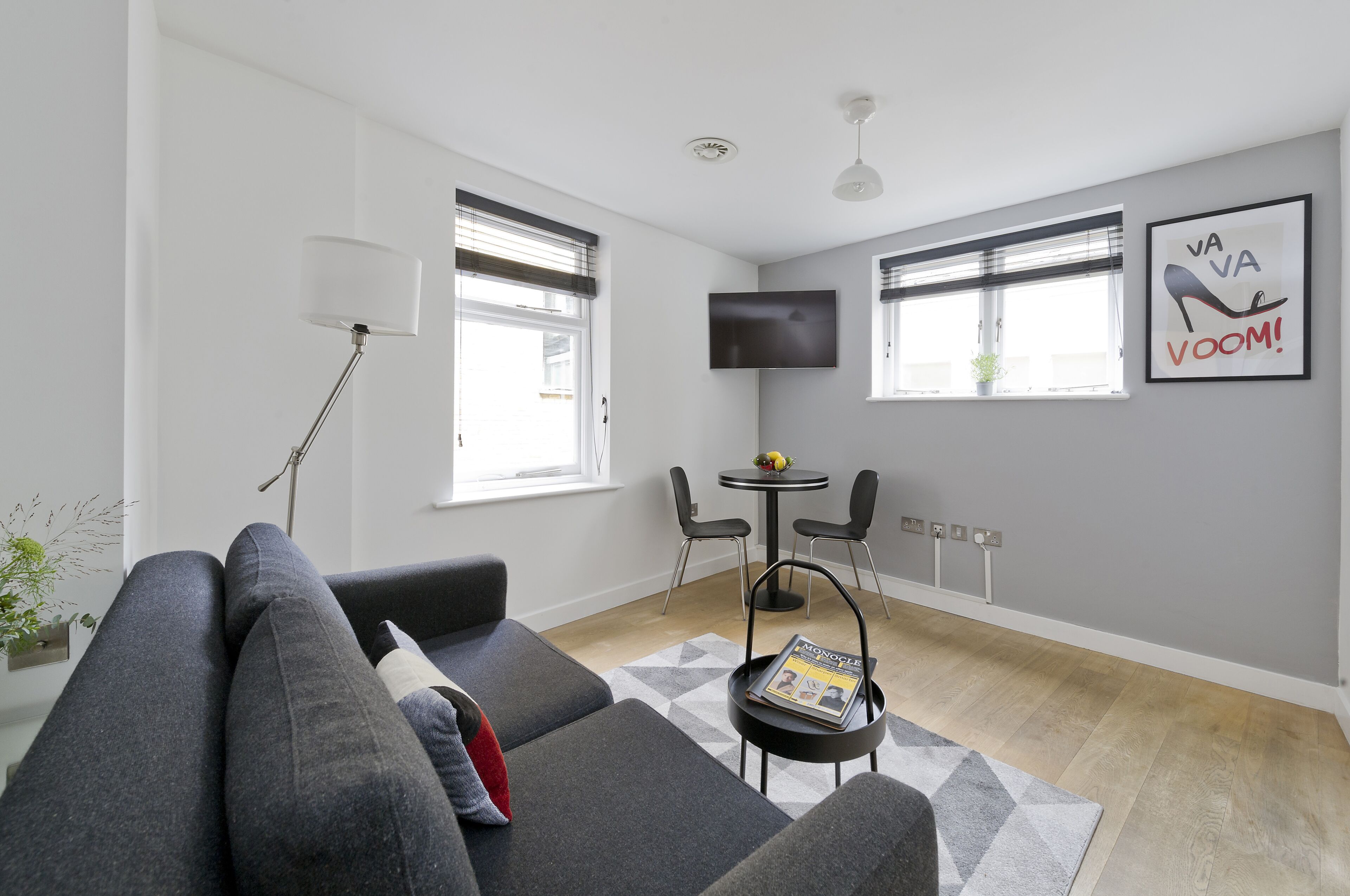 A modern living room with a grey sofa, a small round table with two chairs, a floor lamp, and a flat-screen TV mounted on the wall.