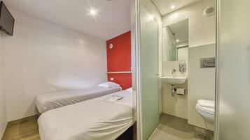 A hotel room with two single beds, a red accent wall, a bathroom with a sink and toilet, and a mirror.