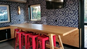 A modern kitchen with a long wooden countertop, red bar stools, a flat-screen TV mounted on the wall, and patterned wallpaper. - Unisotel Le Mans (Le Mans)