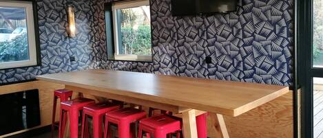 A modern kitchen with a long wooden countertop, red bar stools, a flat-screen TV mounted on the wall, and patterned wallpaper.