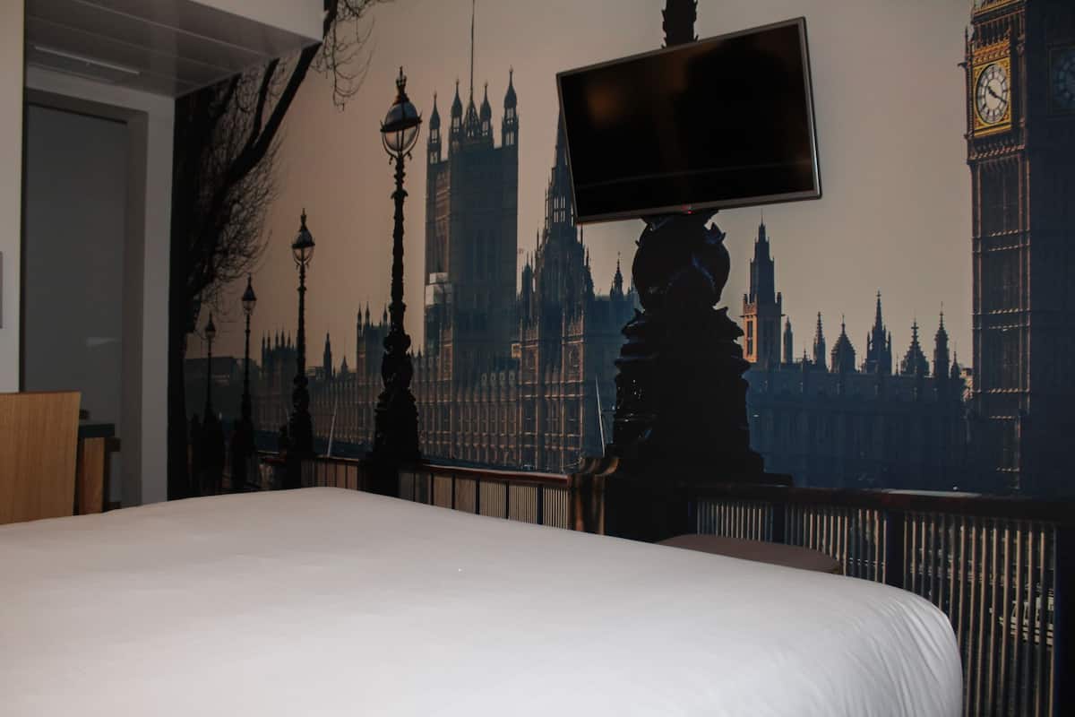 Un dormitorio con una cama grande, un televisor de pantalla plana montado en la pared y un mural con vista a la ciudad al fondo.