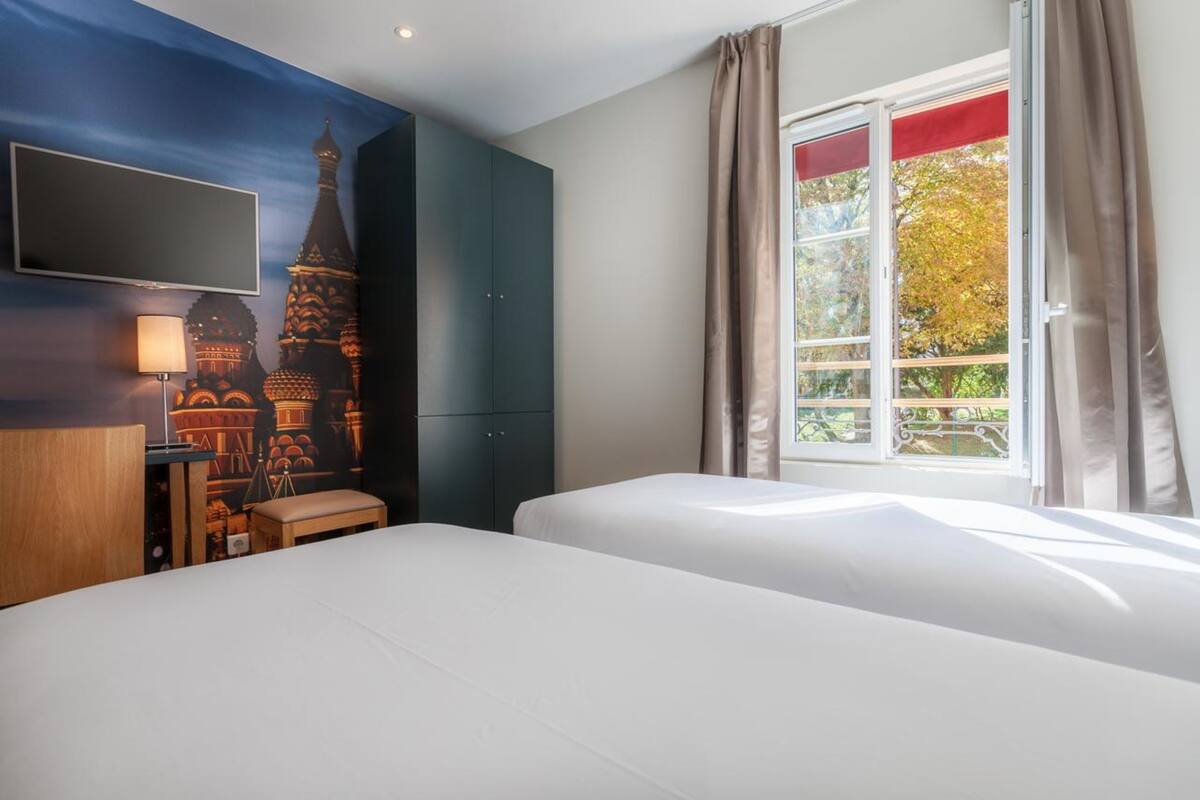 Habitación de hotel con dos camas, un televisor montado en la pared, una ventana con cortinas y un mural de un edificio.