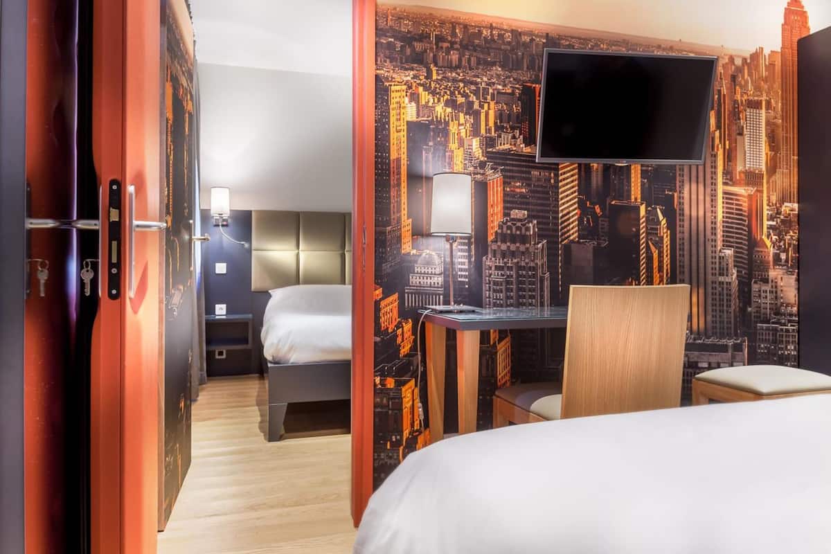 Habitación de hotel con un mural de paisaje urbano, una cama, un escritorio con silla y un televisor montado en la pared.