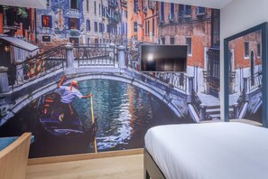 A room with a mural of a canal scene, a bed, and a television mounted on the wall.