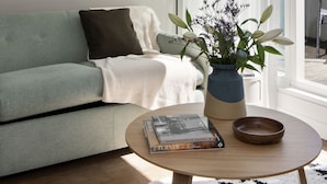 A modern living room with a green sofa, a coffee table with a book and a bowl, a painting on the wall, and a window with sheer curtains. - Cove St Martin’s (London)