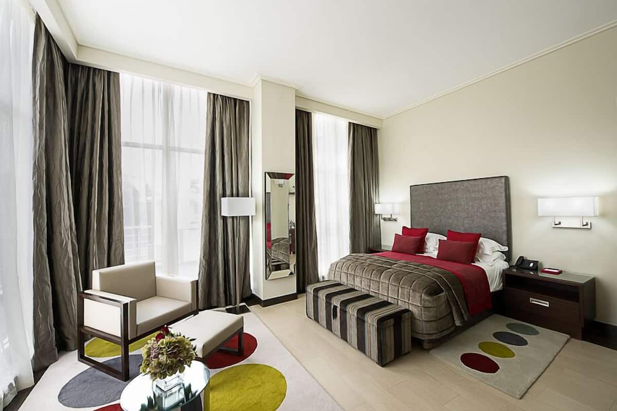 A hotel room with a bed, a chair, a bedside table, a mirror, and a small table with a vase.