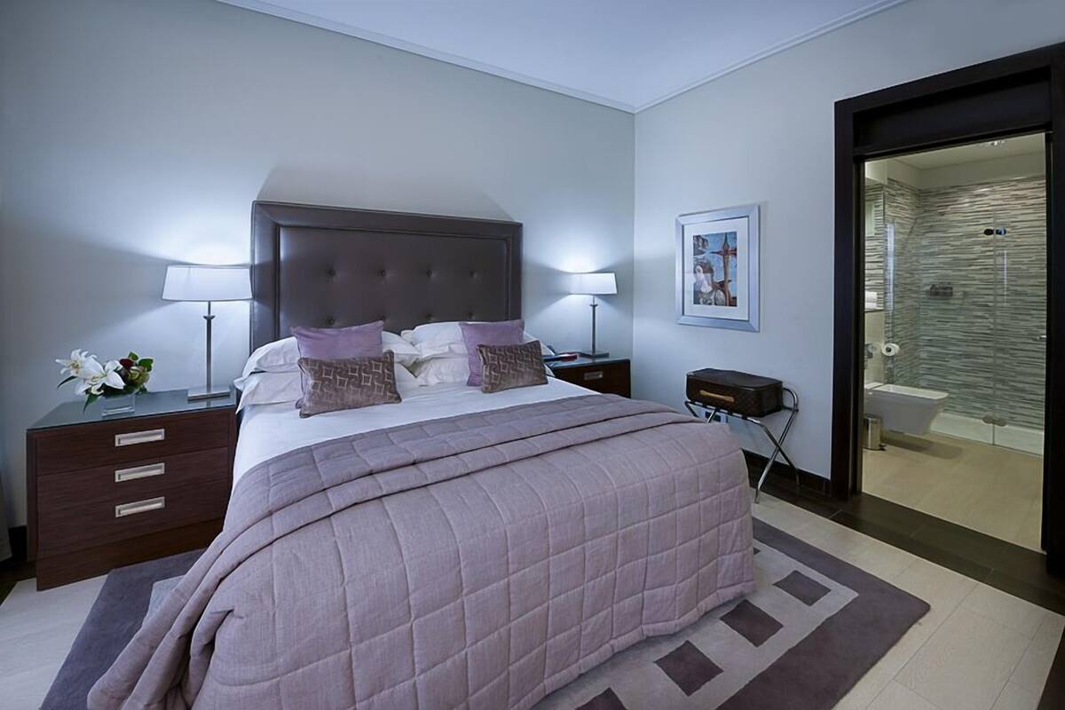 A modern bedroom with a large bed, bedside tables, a nightstand, a framed picture on the wall, and a bathroom visible through an open door.