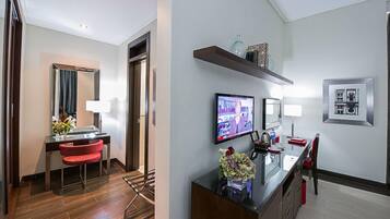 A modern hotel room with a wooden floor, a red chair, a desk with a mirror, a television, and a shelf with decorative items.