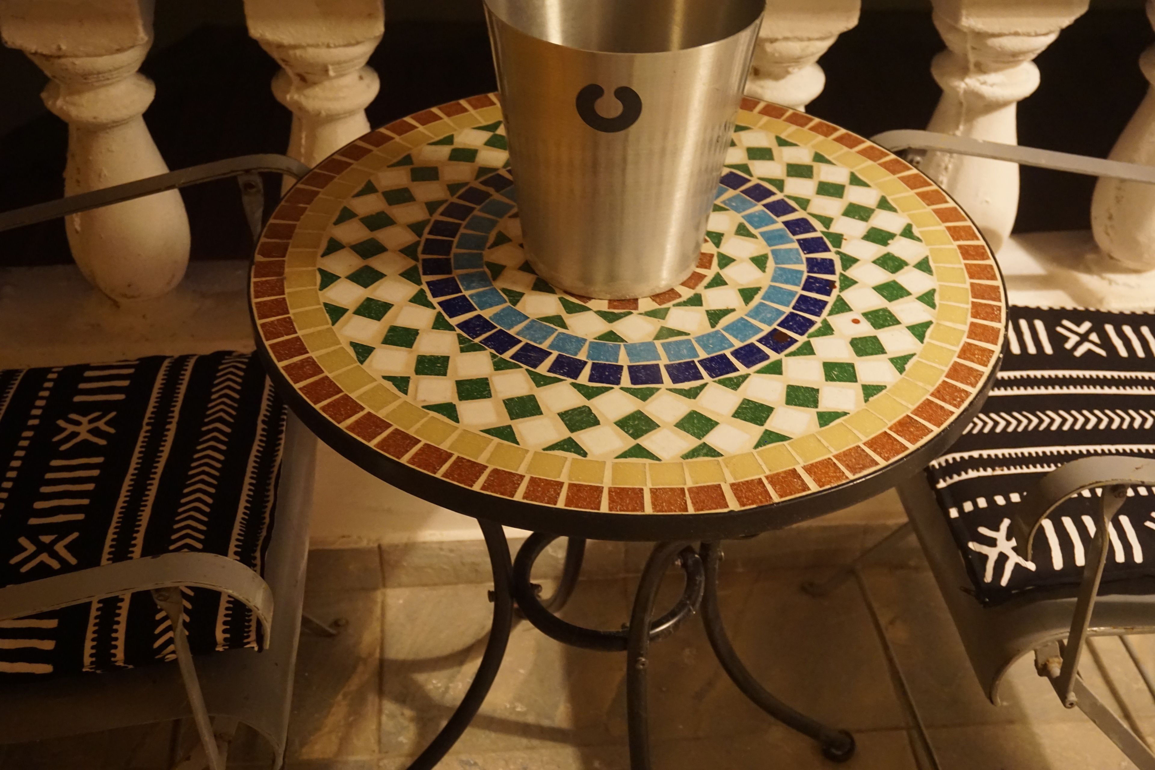 A round table with a mosaic tile top, a metal cup, and a chair with a patterned cushion.